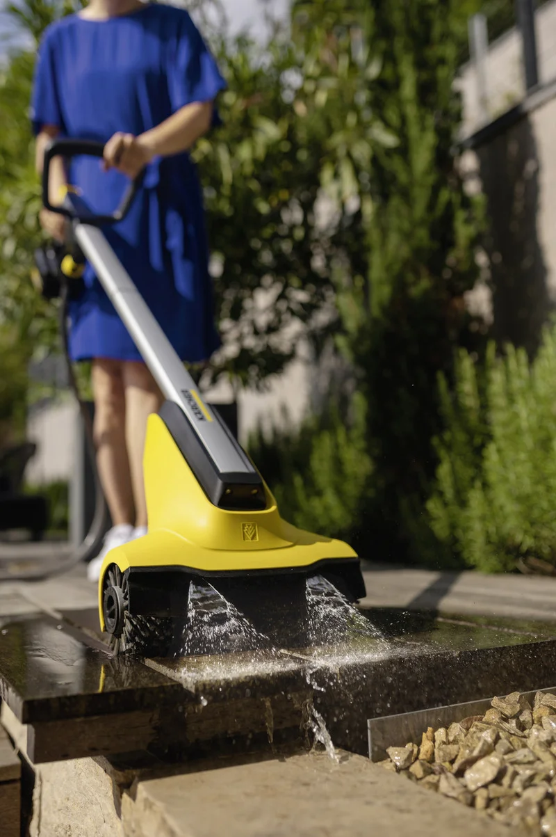 Frau in blauem Kleid reinigt Terrasse mit Kärcher Flächenreiniger. Wasser spritzt auf Steinplatten. Hintergrund: Garten.