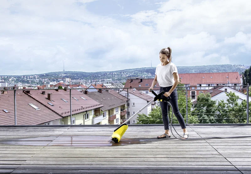 Person reinigt Holzterrasse mit Kärcher Hochdruckreiniger, im Hintergrund Stadtlandschaft mit Hügeln und Häusern.