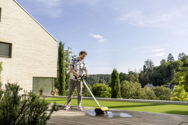 Person reinigt Terrasse mit Kärcher Bodenreiniger. Moderne Hausfassade, grüne Landschaft und blauer Himmel im Hintergrund.