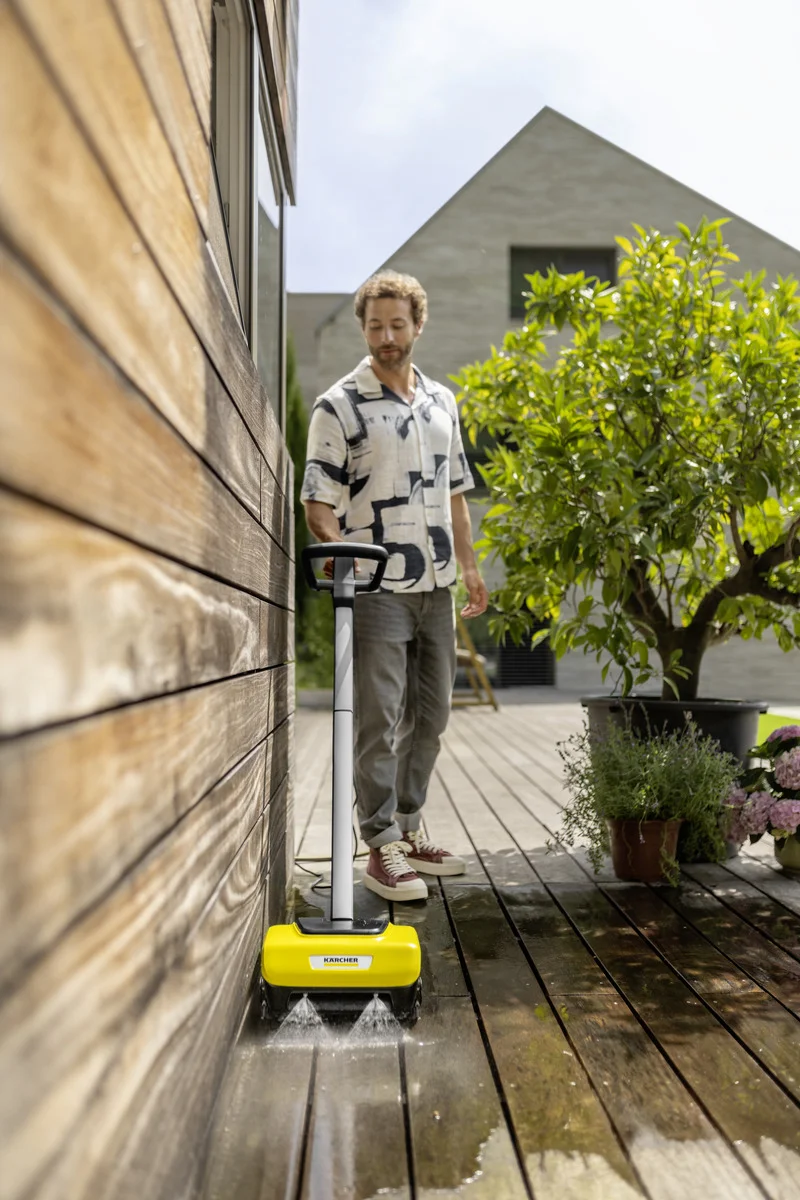 Person reinigt Holzterrasse mit Kärcher Bodenreiniger. Im Hintergrund ein Baum und ein modernes Gebäude.