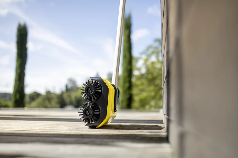 Kärcher Kehrmaschine auf Holzterrasse, zwei rotierende Bürsten, sonniger Tag, Bäume im Hintergrund.