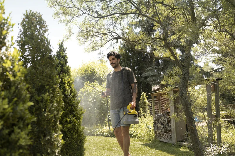 Mann sprüht Pflanzen im Garten mit einem Kärcher Drucksprüher. Umgeben von Bäumen und Sträuchern, sonniges Wetter.