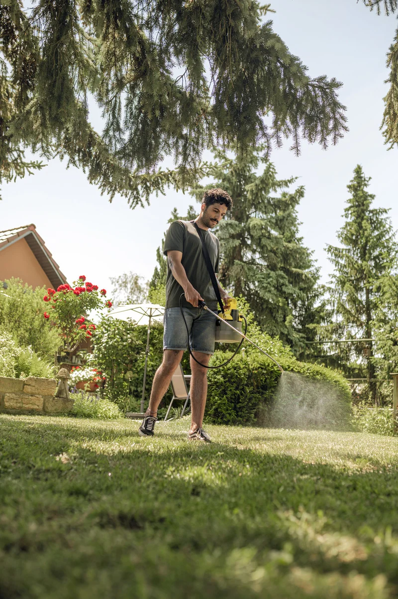 Person sprüht Wasser auf Rasen mit Kärcher Hochdruckreiniger, umgeben von Bäumen und Blumen im Garten.