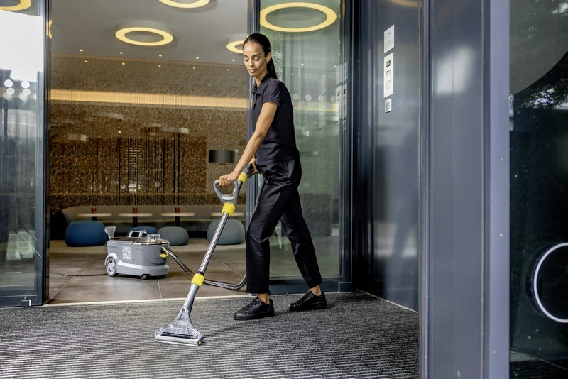 Person reinigt Teppichboden mit Kärcher Teppichreiniger in modernem Innenraum mit Sitzgelegenheiten und stilvoller Beleuchtung.