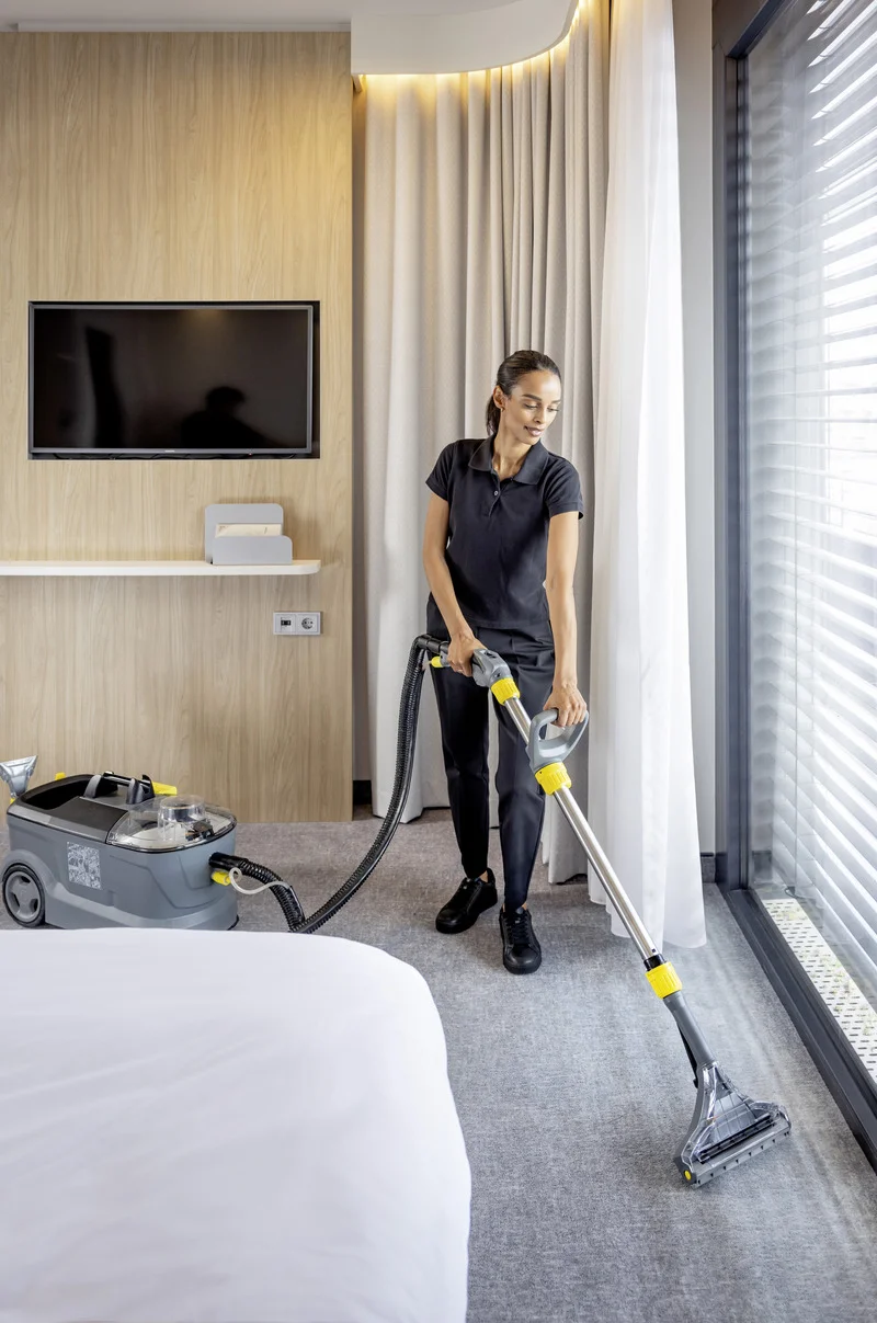Person reinigt Teppichboden in einem modernen Zimmer mit einem Kärcher Teppichreiniger. Im Hintergrund ein Fernseher und große Fenster.