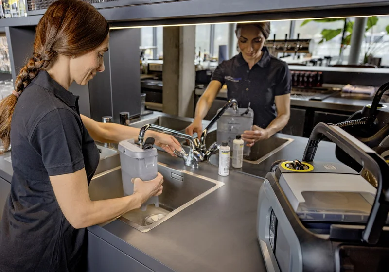 Zwei Personen füllen einen Behälter an einem Wasserhahn in einer modernen Küche. Ein Kärcher-Gerät steht auf der Arbeitsfläche.