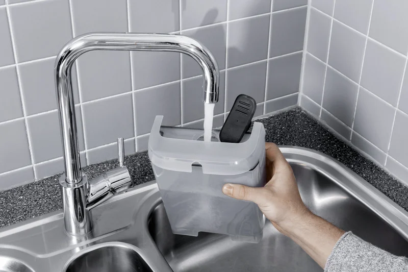 Hand hält Kärcher Wasserbehälter unter fließendem Wasserhahn in einer Edelstahlspüle mit grauen Fliesen im Hintergrund.