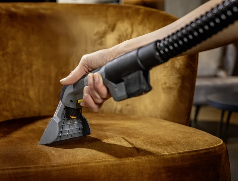 Hand hält Kärcher Polsterdüse, reinigt braunen Sessel in einem Wohnzimmer. Hintergrund unscharf mit Tisch und Sofa.