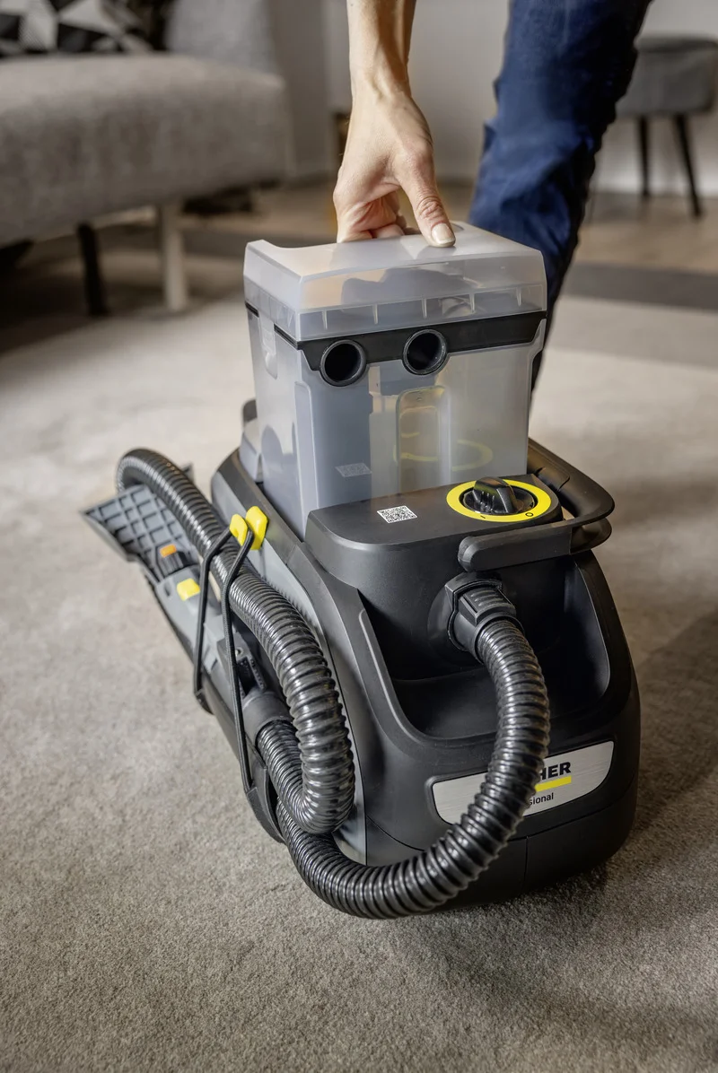 Hand hebt den Wasserbehälter eines Kärcher Teppichreinigers in einem Wohnzimmer an. Gerät steht auf Teppichboden.