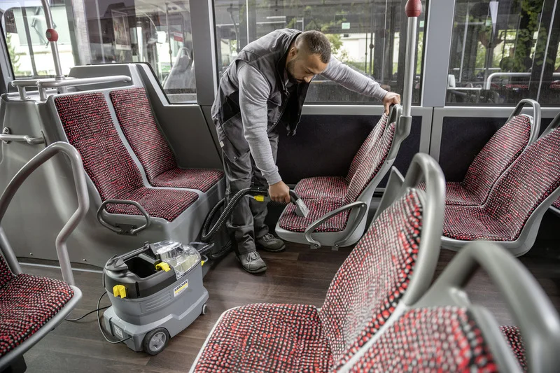 Person reinigt Bussitz mit Kärcher Sprühextraktionsgerät. Sitzbezüge sind rot-schwarz gemustert. Innenraum eines Busses.