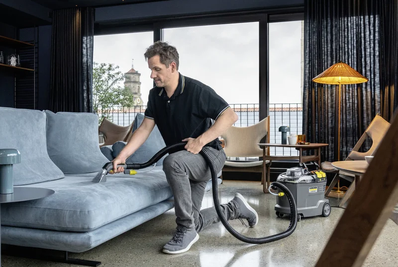 Mann reinigt Sofa mit Kärcher Nass-/Trockensauger in modernem Wohnzimmer mit großen Fenstern und stilvoller Einrichtung.