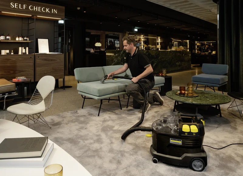 Mann reinigt Sofa mit Kärcher Nasssauger in modernem Hotel-Lobby. Umgebung mit stilvollen Möbeln und "Self Check In"-Bereich.