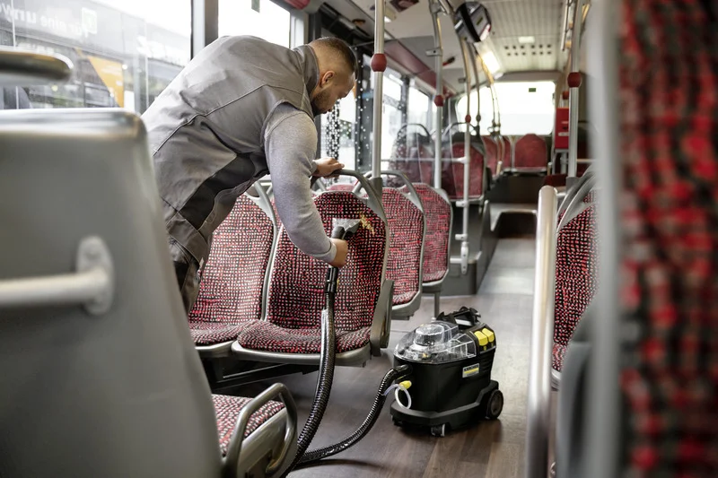 Person reinigt Bussitze mit einem Kärcher Nass-/Trockensauger. Der Bus ist leer und die Sitze haben ein rot-schwarzes Muster.