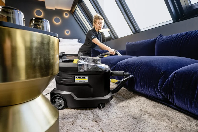 Person reinigt blaue Couch mit Kärcher Nasssauger in modernem Raum mit Dachfenstern und stilvoller Beleuchtung.