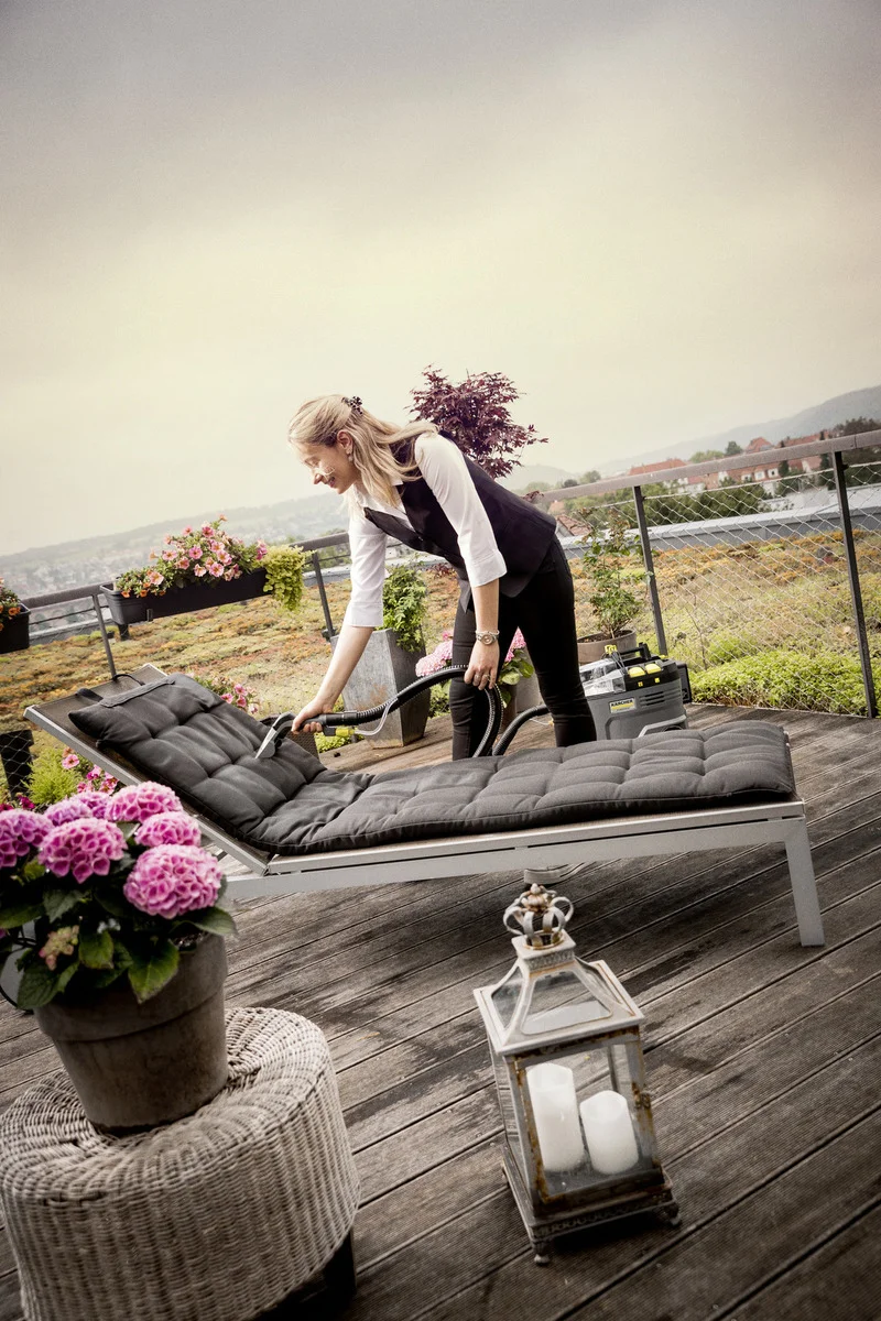Frau reinigt eine Liege auf einer Terrasse mit einem Kärcher Nass-Trockensauger. Im Hintergrund blühende Pflanzen und eine Laterne.