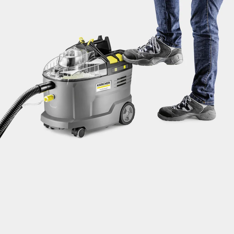 Person in Jeans und Wanderschuhen bedient Kärcher Nass-/Trockensauger mit Fuß. Gerät auf Rollen, Schlauchanschluss sichtbar.