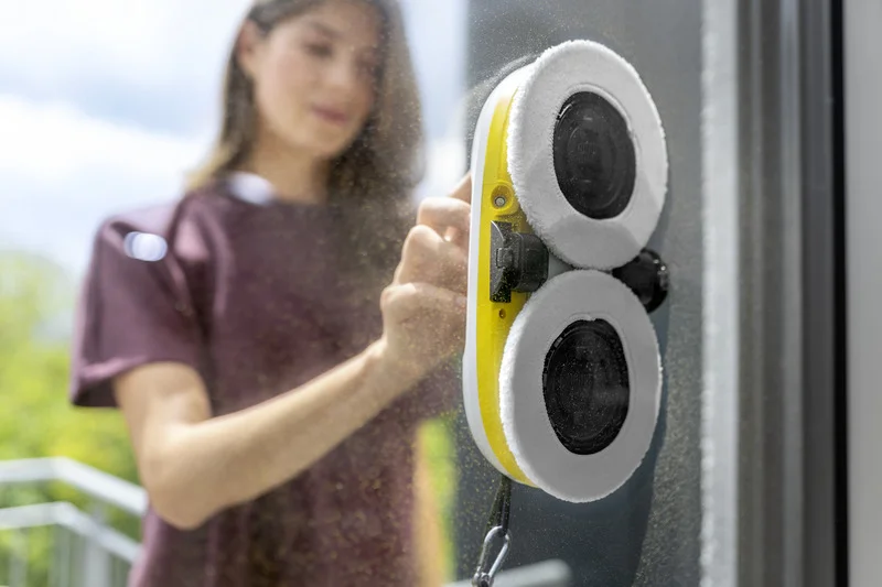 Kärcher Fensterreinigungsgerät an einer Glasscheibe, im Hintergrund verschwommene Person in lila Shirt. Außenbereich mit grünem Hintergrund.