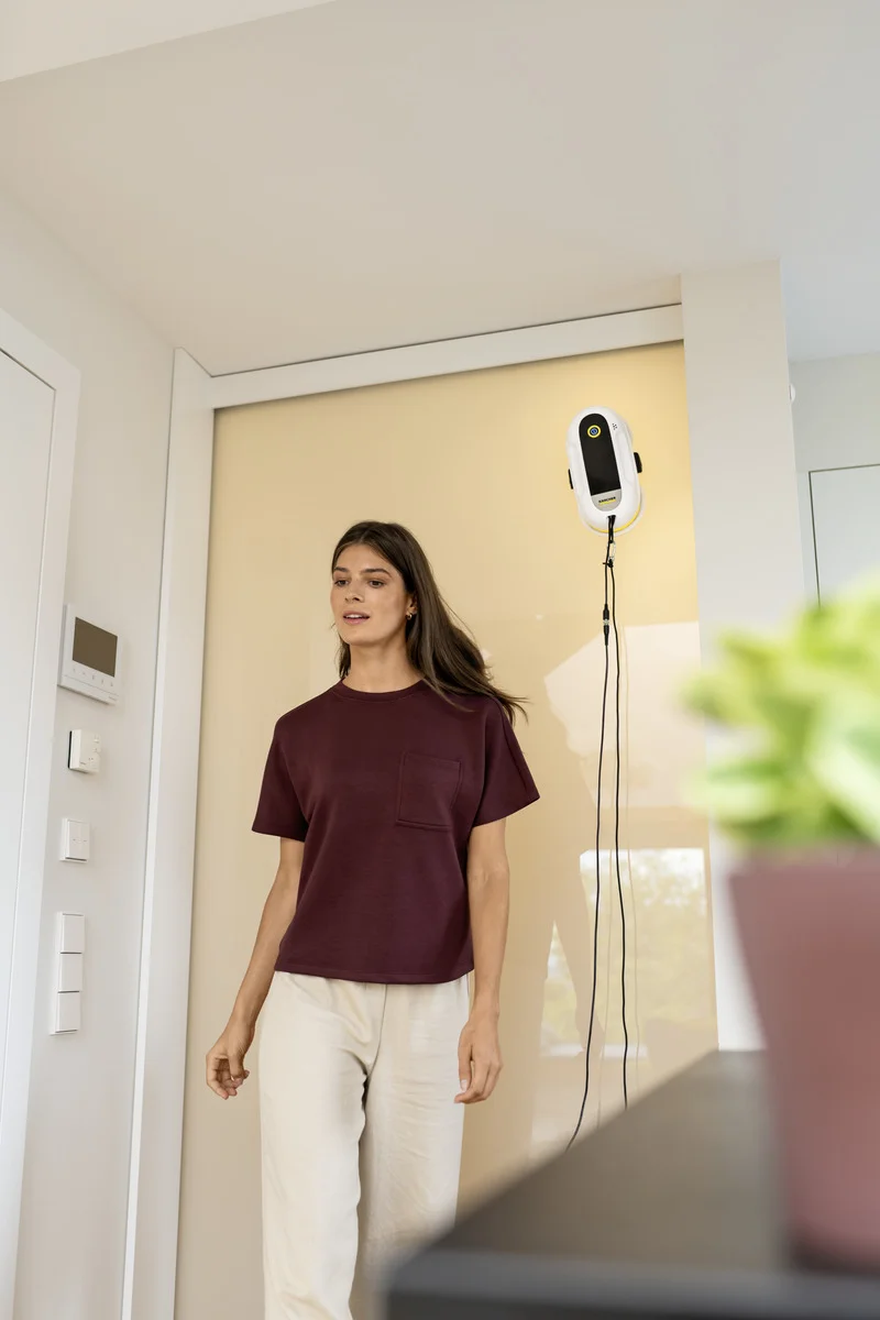Frau in rotem T-Shirt und weißer Hose steht in einem modernen Raum. An der Wand hängt ein Kärcher Fensterreiniger.