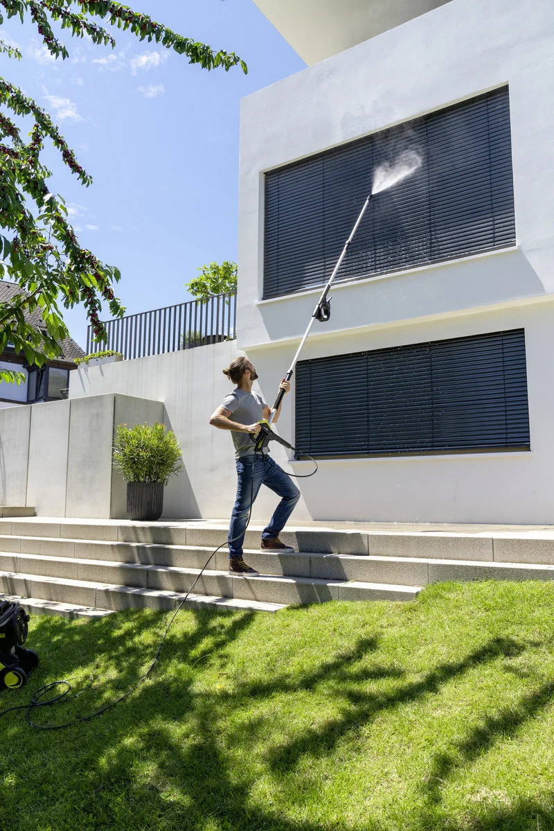 Mann reinigt mit Kärcher Hochdruckreiniger die Fassade eines modernen Hauses. Er steht auf einer Treppe im Garten.