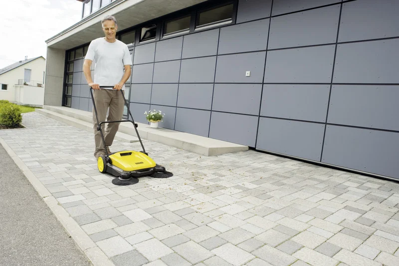 Mann reinigt gepflasterte Einfahrt mit einem gelben Kärcher Kehrmaschine vor einem modernen Gebäude.