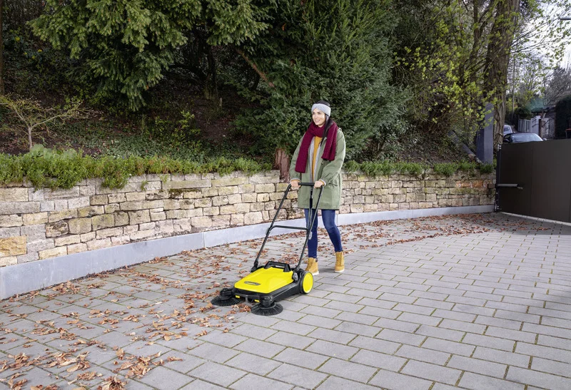 Person nutzt Kärcher Kehrmaschine auf gepflastertem Weg, um Herbstblätter zu entfernen. Hintergrund: Steinmauer und grüne Büsche.