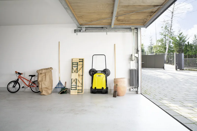 Garage mit rotem Fahrrad, Laubsack, Besen, Kärcher Kehrmaschine, Klapptisch, Blumentopf und Schaufel. Geöffnete Garagentür mit Blick nach draußen.