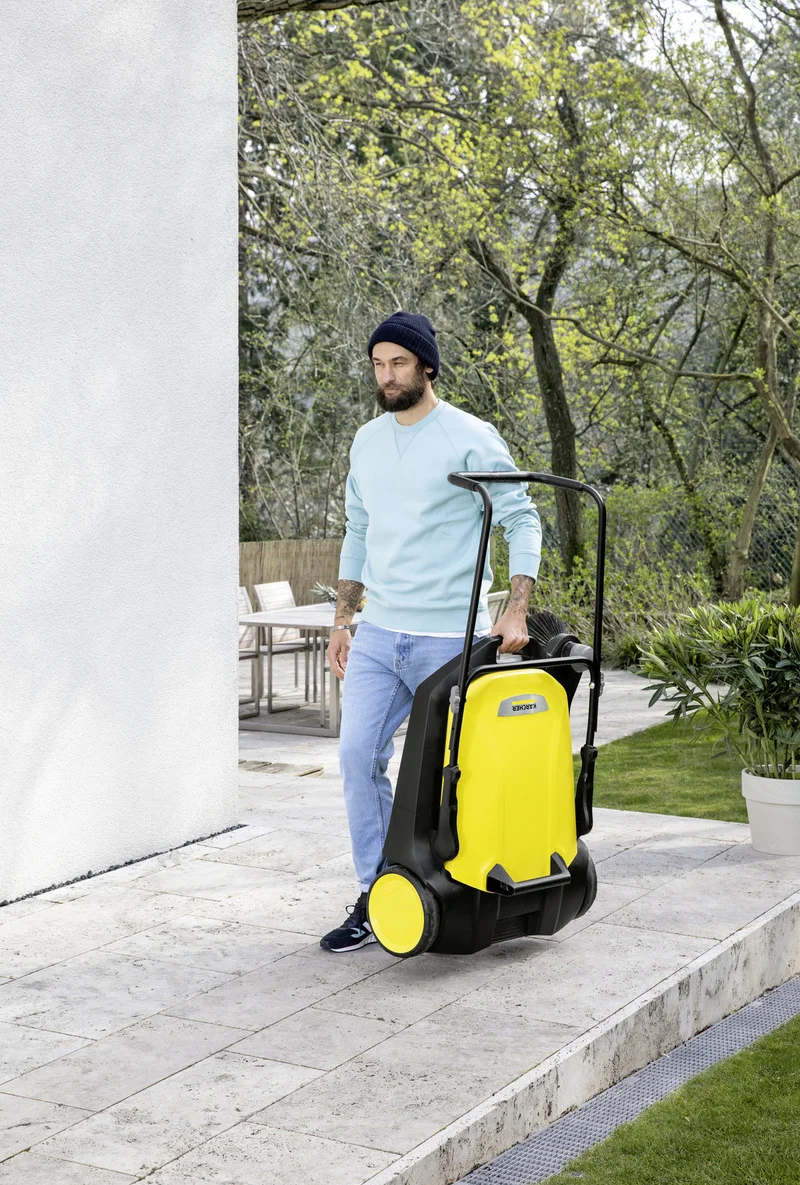 Person im Freien mit einem Kärcher Kehrmaschine auf einer Terrasse, umgeben von Bäumen und Gartenmöbeln.