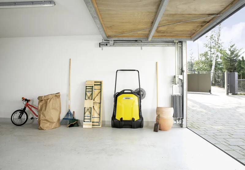Garage mit Kärcher Kehrmaschine, rotem Kinderfahrrad, Laubrechen, Klapptisch, Blumentopf und Gartenschaufel. Offenes Garagentor.