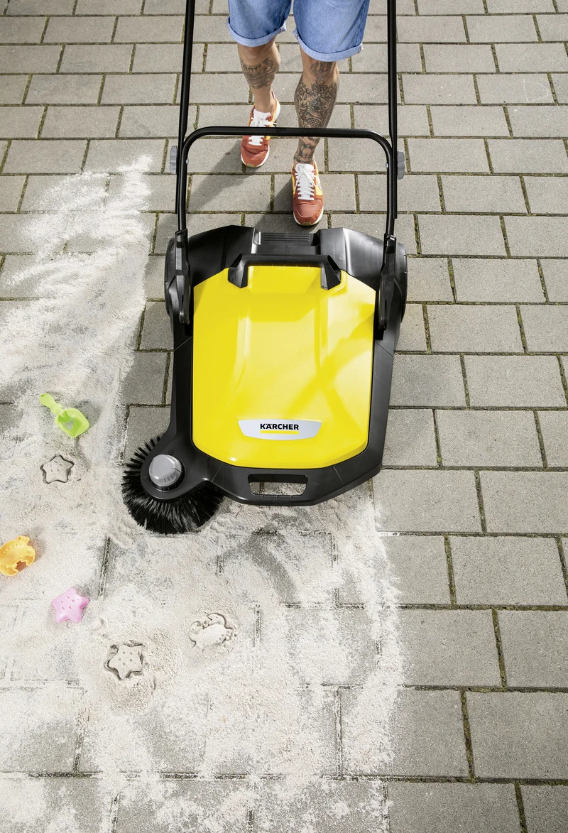 Person mit tätowierten Beinen und roten Schuhen schiebt einen Kärcher Kehrmaschine über sandige Pflastersteine.