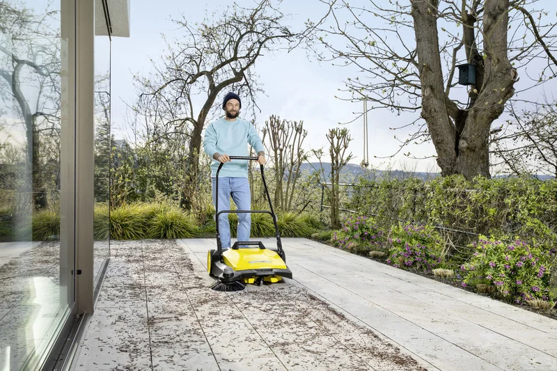 Person reinigt Terrasse mit Kärcher Kehrmaschine, umgeben von Bäumen und blühenden Pflanzen im Freien.
