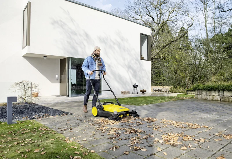 Person nutzt Kärcher Kehrmaschine auf gepflastertem Weg vor modernem Haus, um Herbstblätter zu entfernen. Garten und Bäume im Hintergrund.
