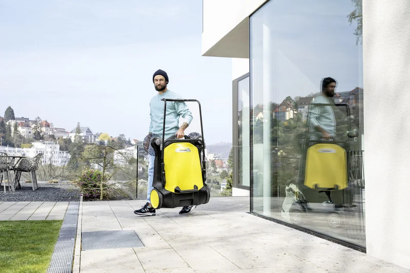 Person im Freien mit einem Kärcher Kehrmaschine, neben einem modernen Gebäude mit Glasfassade, im Hintergrund eine städtische Landschaft.