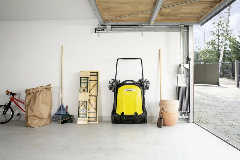 Kärcher Kehrmaschine in Garage neben Fahrrad, Laubsack, Rechen, Klapptisch, Blumentopf und Schaufel. Geöffnete Garagentür zeigt Außensicht.