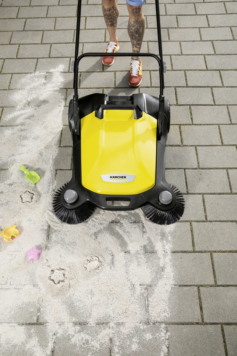 Kärcher Kehrmaschine auf Pflastersteinen, Sand und Spielzeugformen, tätowierte Beine und rote Schuhe im Hintergrund.