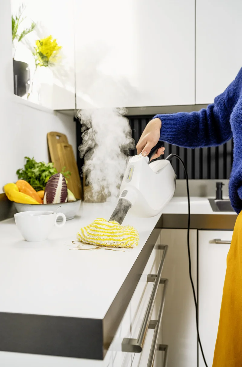 Person reinigt Küchenarbeitsplatte mit einem Kärcher Dampfreiniger. Gemüse und Tasse stehen daneben.