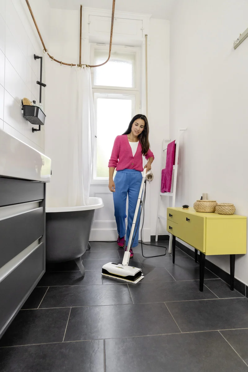 Frau reinigt Badezimmerboden mit einem Kärcher Dampfreiniger. Umgebung: Badewanne, gelber Schrank, Handtücher auf Leiter.