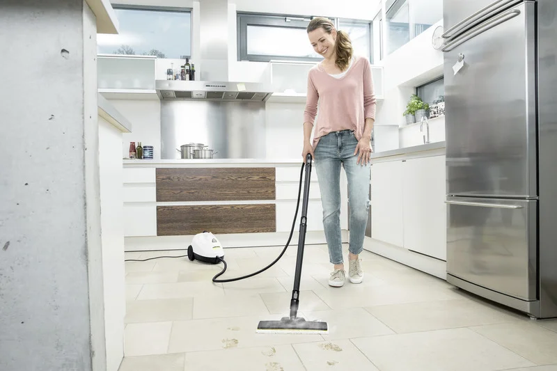 Frau reinigt Küchenboden mit einem Kärcher Dampfreiniger. Moderne Küche mit Edelstahlgeräten und großen Fenstern.
