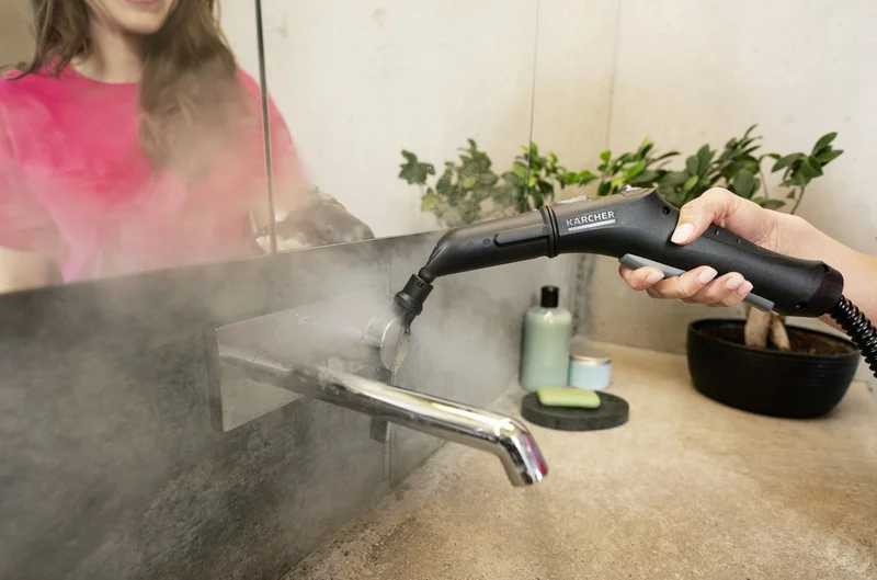 Hand hält Kärcher Dampfreiniger, reinigt Wasserhahn im Badezimmer. Frau im Hintergrund, Pflanzen und Seife auf dem Waschbecken.