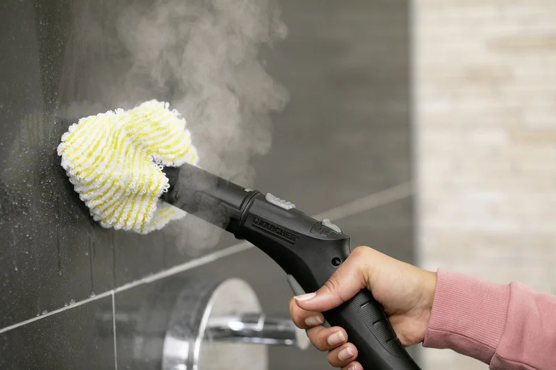 Hand hält Kärcher Dampfreiniger, der mit einem gelben Reinigungstuch eine dunkle Fliese reinigt.