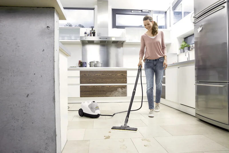 Frau reinigt Küchenboden mit Kärcher Dampfreiniger. Moderne Küche mit Edelstahlgeräten und großen Fenstern.
