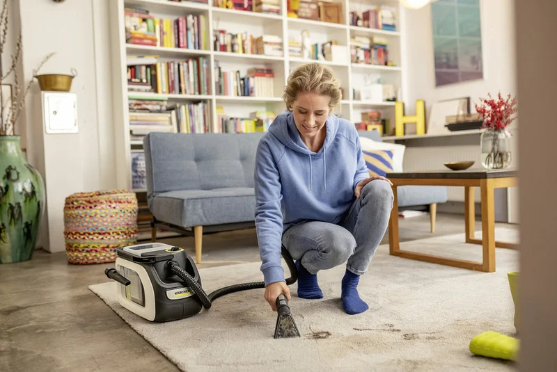 Person reinigt Teppich mit Kärcher Teppichreiniger in gemütlichem Wohnzimmer mit Bücherregal und blauer Couch.