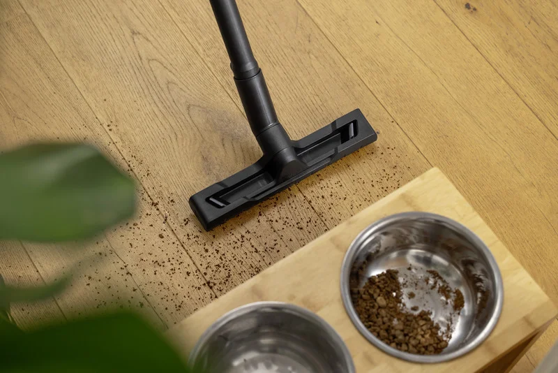Staubsauger entfernt Krümel von Holzboden neben Futterstation mit zwei Edelstahlnäpfen.