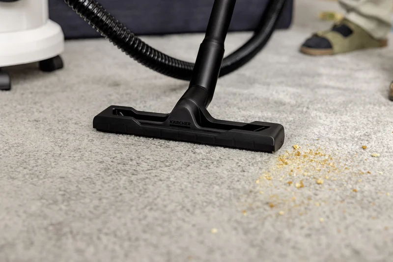 Kärcher Staubsaugerdüse auf Teppich, saugt Krümel auf. Im Hintergrund sind Füße in Sandalen sichtbar.