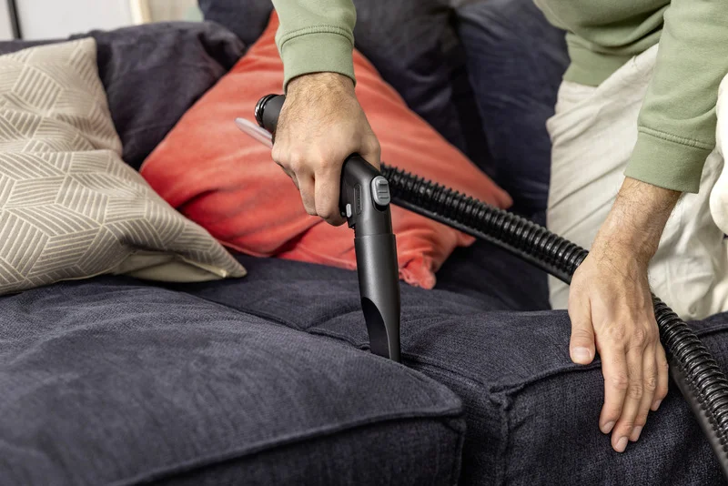 Person reinigt dunkelgraues Sofa mit Kärcher Staubsauger. Zwei Kissen, eines rot, eines mit geometrischem Muster, liegen auf dem Sofa.