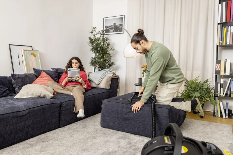 Person reinigt Sofa mit Kärcher Staubsauger, während andere Person auf Sofa liest. Wohnzimmer mit Pflanzen und Bücherregal im Hintergrund.