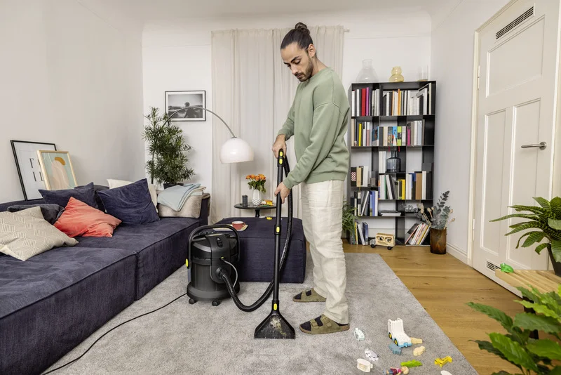 Person reinigt Teppich mit Kärcher Waschsauger in modernem Wohnzimmer. Sofa, Bücherregal und Spielzeug im Hintergrund.