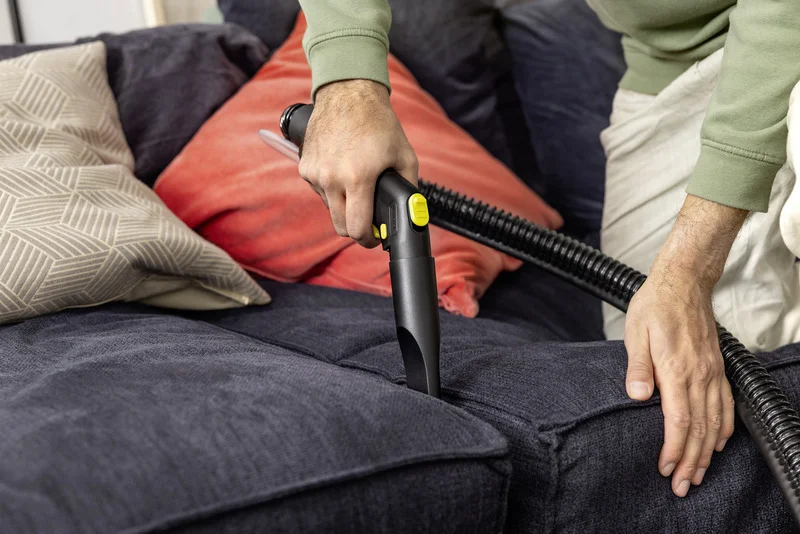 Person reinigt Sofa mit Kärcher Staubsauger. Mehrere Kissen im Hintergrund, darunter ein rotes und ein gemustertes Kissen.
