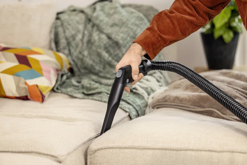 Hand hält Kärcher Staubsaugerdüse, reinigt helles Sofa. Bunte Kissen und grüne Decke im Hintergrund.