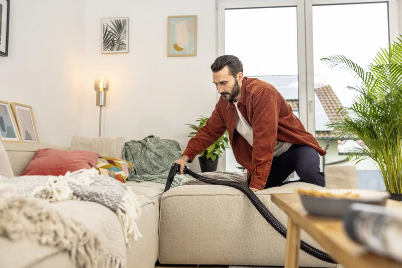 Person reinigt Sofa mit einem Staubsauger in einem gemütlichen Wohnzimmer. Im Hintergrund sind Pflanzen und Wandbilder zu sehen.