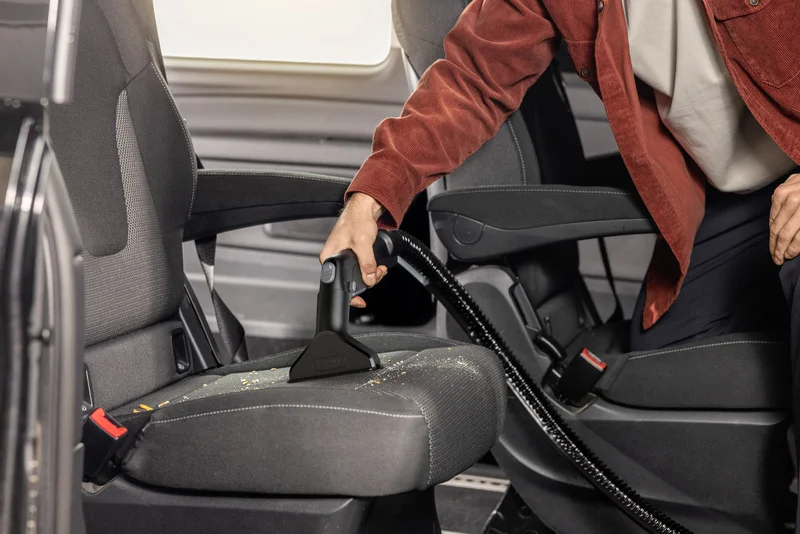 Person reinigt Autositz mit Kärcher Handgerät. Sitz ist mit Krümeln bedeckt. Person trägt rote Jacke und weiße Kleidung.